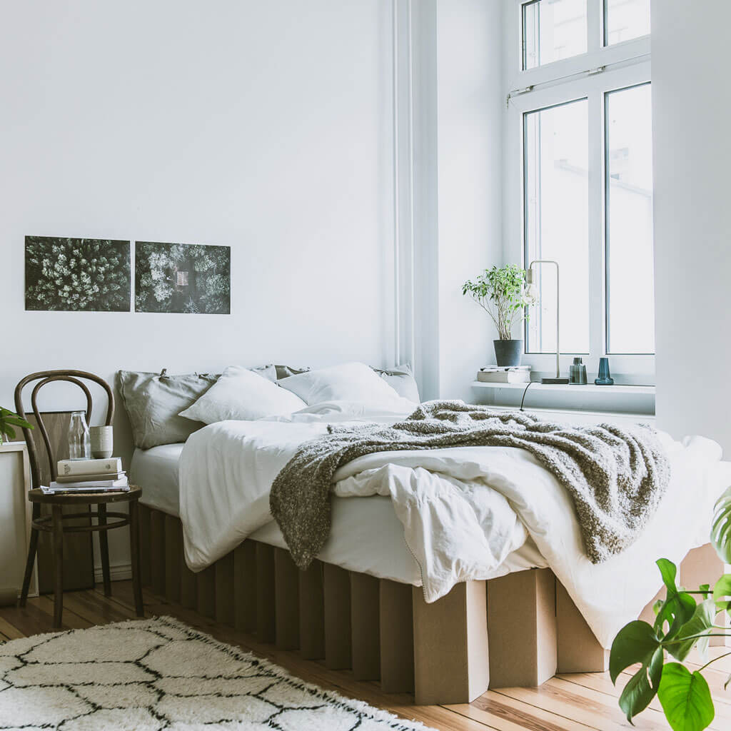 Bett 2.0, a sustainable cardboard bed frame by RIAB USA, surrounded by artworks, plants, and messy bedding in morning light