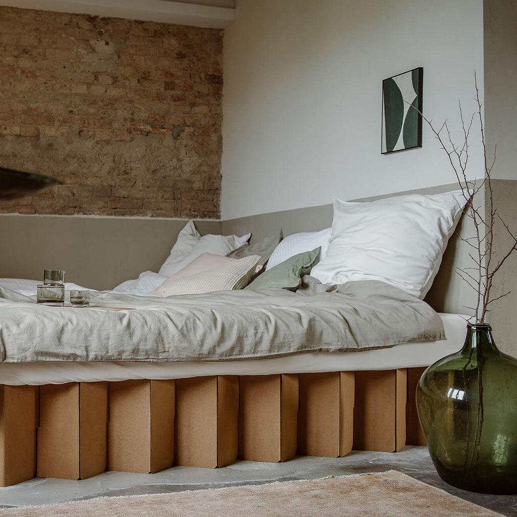 Bett 2.0, a sustainable cardboard bed frame by RIAB USA, against a brick wall with glassware showcasing sturdiness