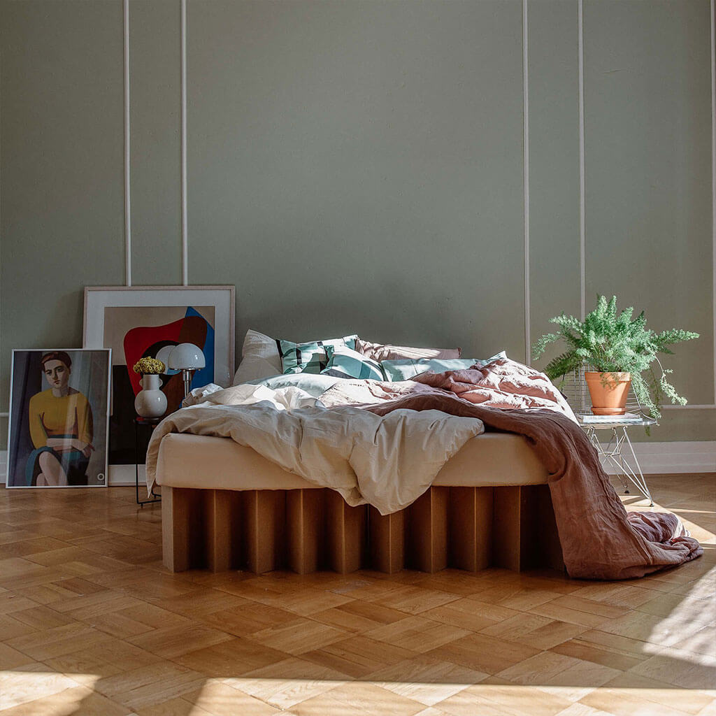 Bett 2.0, a sustainable cardboard bed frame by RIAB USA, surrounded by artwork, plants, and messy bedding in morning light