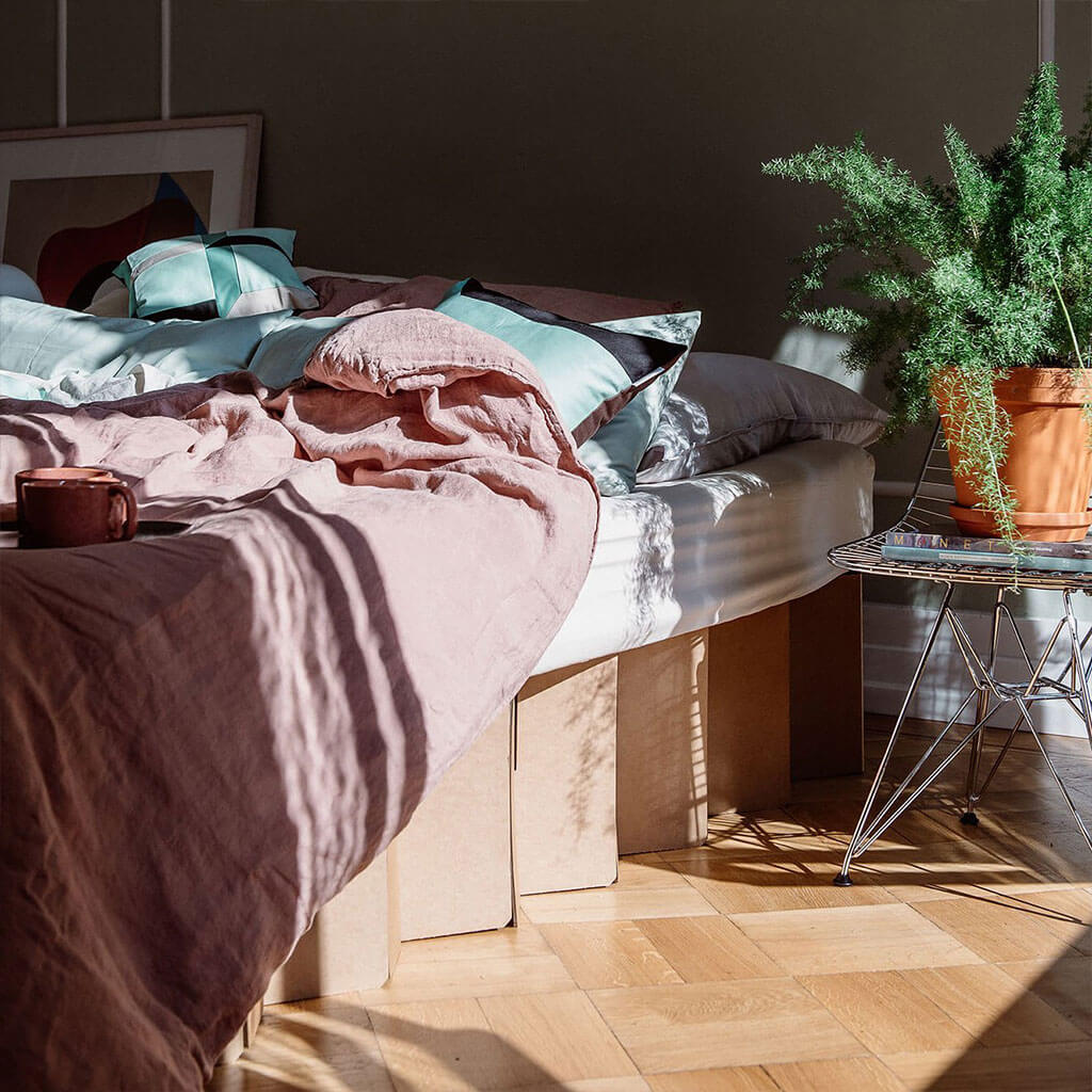 Bett 2.0, a resilient cardboard bed frame by RIAB USA, with messy linens, coffee mug, and sunlight on a plant implying morning